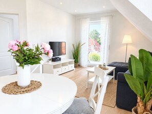 A modern living room with a white TV stand, a dining table with chairs, a black sofa, and a large window with curtains.