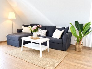 A modern living room with a dark grey sofa, a white coffee table, a woven rug, and a potted plant.