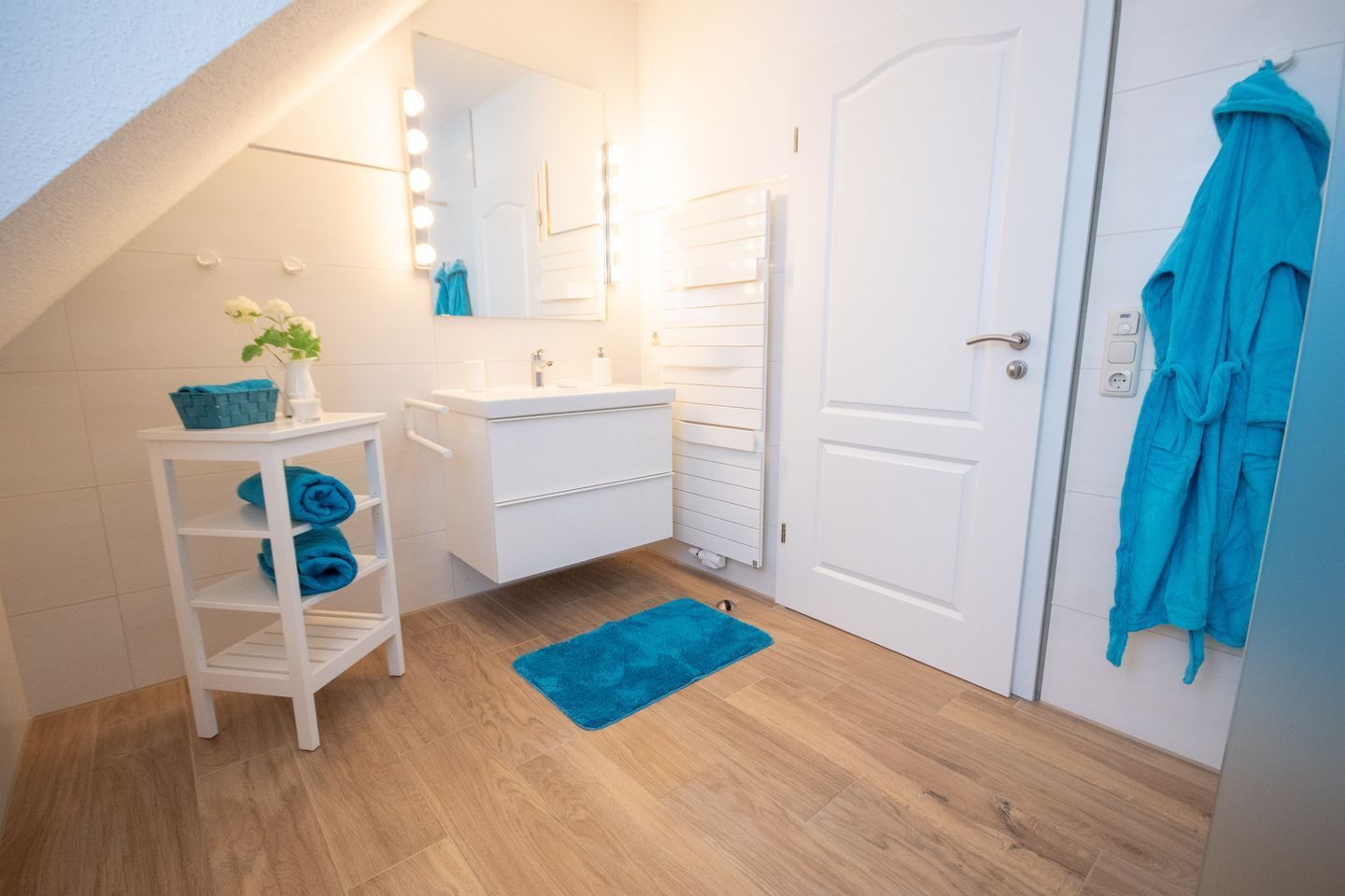 A bathroom with a white vanity, a mirror with lights, a blue towel rack, and a blue rug.