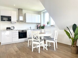 A modern kitchen with white cabinetry, a built-in oven, and a dining area with white chairs and a table. - DS5 - Strandnahe Modernes Ferienapartment für bis zu 4 Personen in Duhnen - Duhner Strand (Cuxhaven)