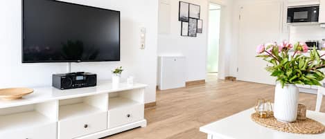 A modern living room with a flat-screen TV, a white entertainment unit, a dining table with a vase of flowers, and a kitchen area in the background.