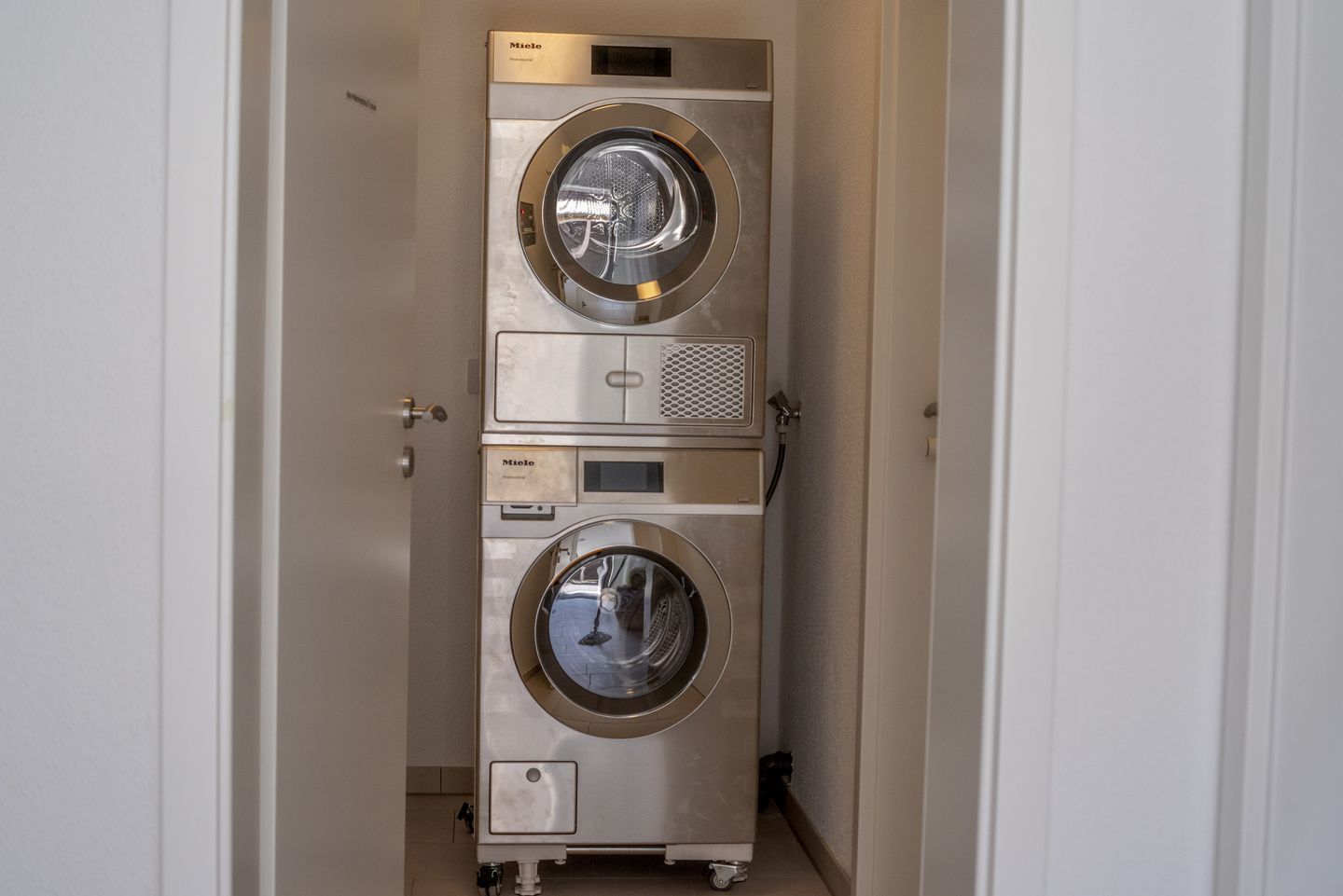 A compact, stainless steel, wall-mounted washer and dryer unit with a control panel and a small storage compartment.