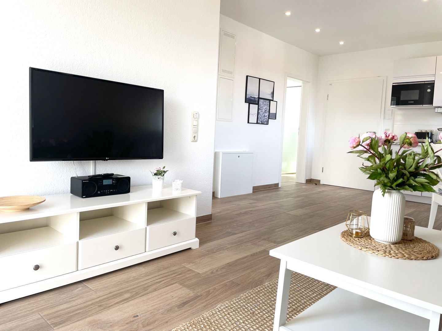 Ein modernes Wohnzimmer mit einem Flachbildfernseher, einer weißen Unterhaltungseinheit, einem Esstisch mit Blumenvase und einer Küche im Hintergrund.