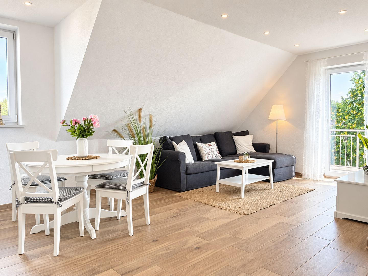 A modern living room with a dark grey sofa, white chairs, a small white table, and a dining table with chairs. There is a large window with a view of greenery outside.