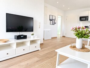Ett modernt vardagsrum med en platt-TV, en vit möbel för underhållningssystemet, ett matbord med en vas med blommor och ett köksområde i bakgrunden.