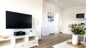 A modern living room with a flat-screen TV, a white entertainment unit, a dining table with a vase of flowers, and a kitchen area in the background.