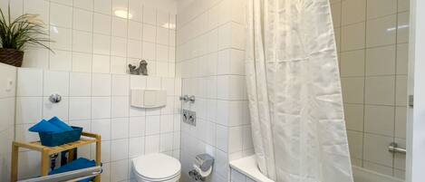 Un baño con inodoro, ducha con cortina, lavabo y un estante pequeño con planta y toallas azules.