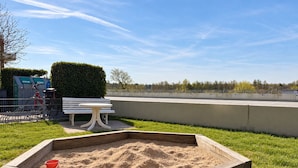 A sandy play area with a bench and a table, surrounded by a fence and a hedge.