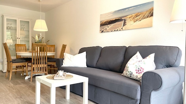 A living room with a grey sofa, white cushions, a small white table, wooden chairs, a glass cabinet, and a beach-themed wall art.