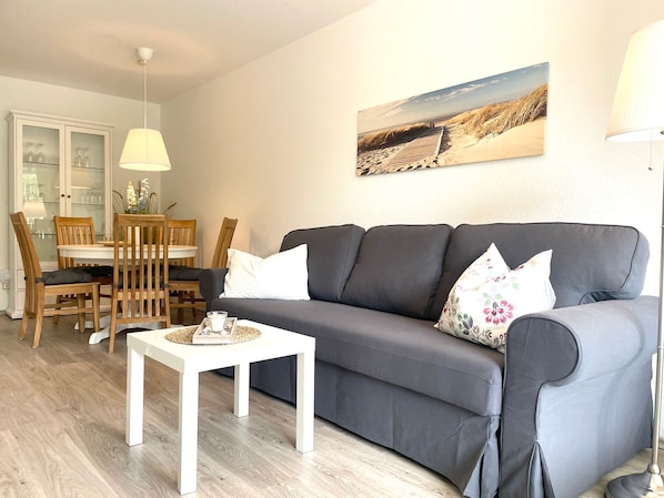 A living room with a grey sofa, white cushions, a small white table, wooden chairs, a glass cabinet, and a beach-themed wall art.