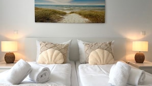 A hotel room with two beds, white linens, a beach-themed wall hanging, and two bedside lamps.