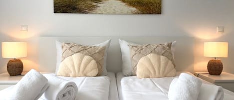 A hotel room with two beds, white linens, a beach-themed wall hanging, and two bedside lamps.