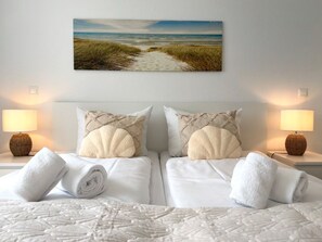 A hotel room with two beds, white linens, a beach-themed wall hanging, and two bedside lamps.