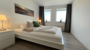 A bedroom with a bed, a nightstand, a lamp, a painting, and two windows with curtains. - A1.4 - Exclusive vacation apartment with balcony, close to the beach for up to 7 people - (Cuxhaven)