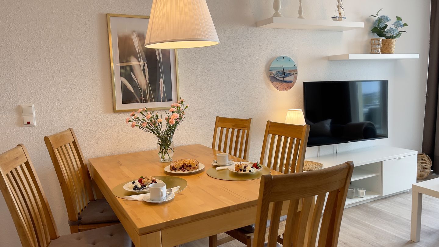 Un comedor con una mesa de madera, sillas, un jarrón de flores y un televisor sobre un soporte blanco.