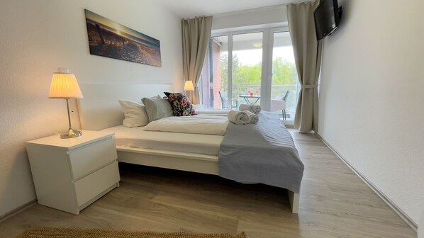 Un dormitorio con cama, mesita de noche, lámpara, cuadro, televisor y balcón con silla.