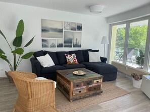 Una sala de estar con un sofá oscuro, un sillón de mimbre, una mesa de centro de madera y un ventanal con vistas a zonas verdes.