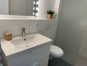 Une salle de bain avec un lavabo blanc, un miroir avec éclairage intégré, un distributeur de savon en bois, une plante en pot et des toilettes.
