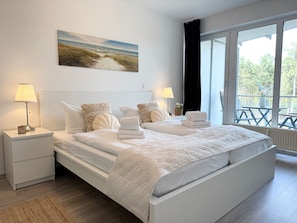 Una cama doble con cabecera blanca, dos mesitas de noche con lámparas, un cuadro de una playa y un gran ventanal con vistas a árboles y una terraza.