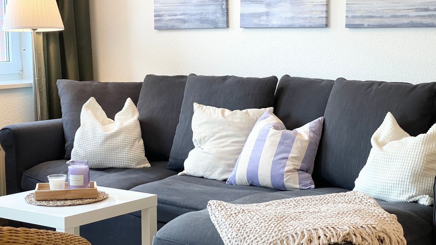 Ein graues Sofa mit gestreiften und strukturierten Kissen, ein weißer Nachttisch mit Kerze und Tablett sowie eine gewebte Tagesdecke.