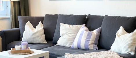 Un sofá gris con almohadones a rayas y con textura, una mesita auxiliar blanca con una vela y una bandeja, y una manta tejida.