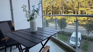 Un balcón con mesa y sillas, un jarrón con flores y vista a una calle con una camioneta blanca.