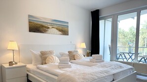 Una cama doble con cabecera blanca, dos mesitas de noche con lámparas, un cuadro de una playa y un gran ventanal con vistas a árboles y una terraza.