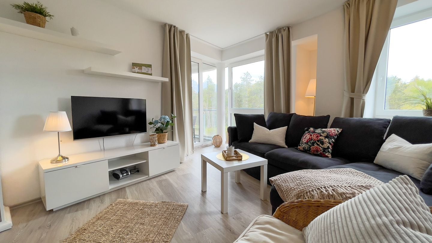 A modern living room with a flat-screen TV, a white entertainment unit, a black sofa with floral cushions, a small white coffee table, and a woven rug.