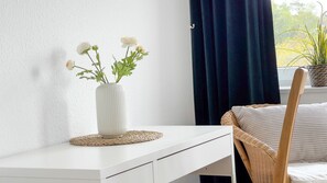 A white bedside table with a white vase containing white flowers, a wicker chair with a striped cushion, and a window with a dark curtain. - A2.3 - Comfortable beachfront apartment for 7 people - House (Cuxhaven)