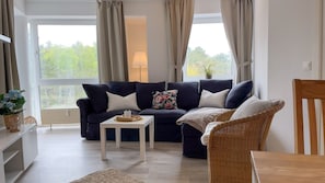 A living room with a dark blue sofa, a white side table, a wicker chair, and a view of trees through large windows.