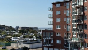 A multi-story residential building with balconies, a parking area, and a view of the sea.
