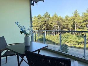 Un balcón con mesa y sillas, un florero con flores y vistas a una zona boscosa.