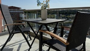 A balcony with a table and two chairs, a vase with flowers, and a view of a parking area and buildings.