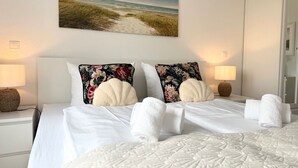 A hotel room with a bed, two pillows, a nightstand with a lamp, a wall-mounted painting of a beach, and a wardrobe.