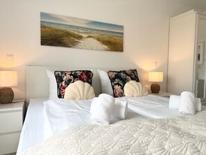 A hotel room with a bed, two pillows, a nightstand with a lamp, a wall-mounted painting of a beach, and a wardrobe. - A3.1 - Modern, cozy vacation apartment, close to the beach for up to 6 people - House (Cuxhaven)