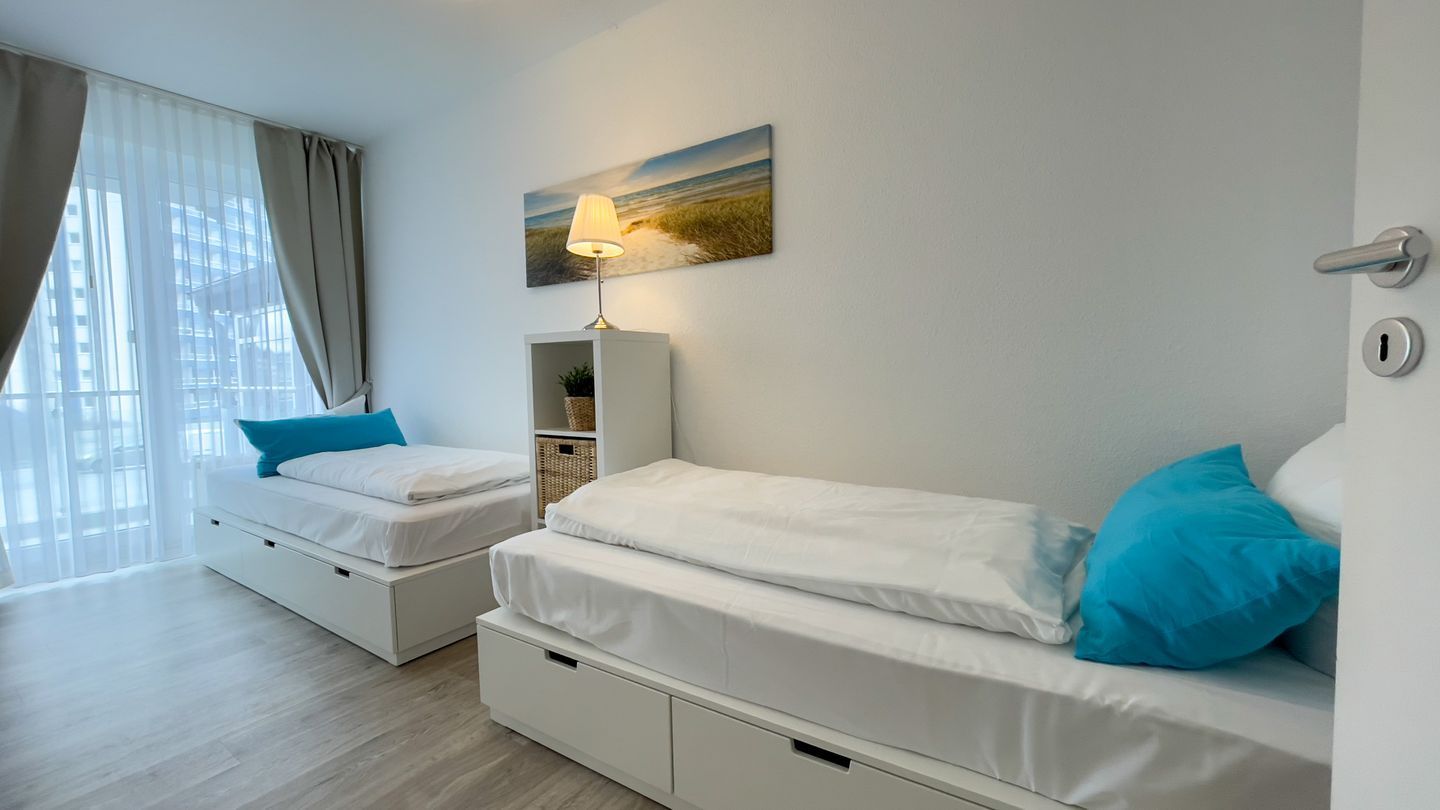 Una camera d'albergo con due letti, un comodino con lampada, un quadro appeso al muro e una finestra con tende.