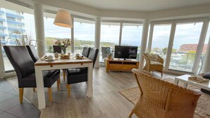 A modern living room with a dining table, chairs, a TV, and large windows offering a city view.