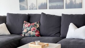 Ein graues Sofa mit blumigen und weißen Kissen, ein Holztablett mit einem Glas und einer Kerze sowie vier gerahmte Bilder an der Wand.