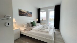 A bedroom with a bed, a nightstand, a lamp, a painting, and a window with curtains.