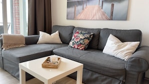 Un canapé d’angle gris avec des coussins blancs et à motifs floraux, une table basse blanche avec une bougie et un plateau, et un tableau représentant un quai accroché au mur.