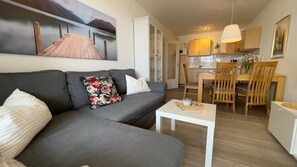 Un salon avec un canapé gris, une table basse blanche et une table à manger en bois avec des chaises. Un tableau représentant un quai et des montagnes est accroché au mur.