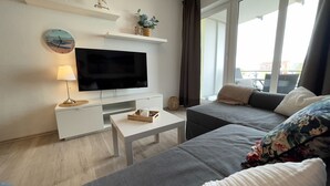 Un salon moderne avec un téléviseur à écran plat, une table basse blanche, un canapé gris et une fenêtre donnant sur un beau paysage.
