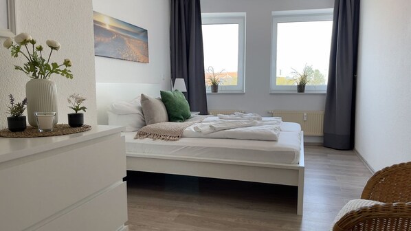 Une chambre à coucher avec un lit, une table de chevet, un vase de fleurs, un tableau et une fenêtre avec des rideaux.