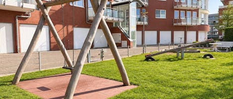 A playground with swings and a seesaw in a grassy area with a brick building in the background.