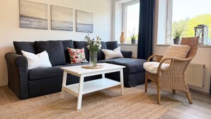 Een moderne woonkamer met een donkergrijze bank, een witte salontafel, een rieten fauteuil en kunst aan de muur.