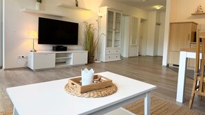 Een moderne woonkamer met een flatscreen televisie, witte meubels, een houten tafel met decoratieve objecten en een keuken in de achtergrond.