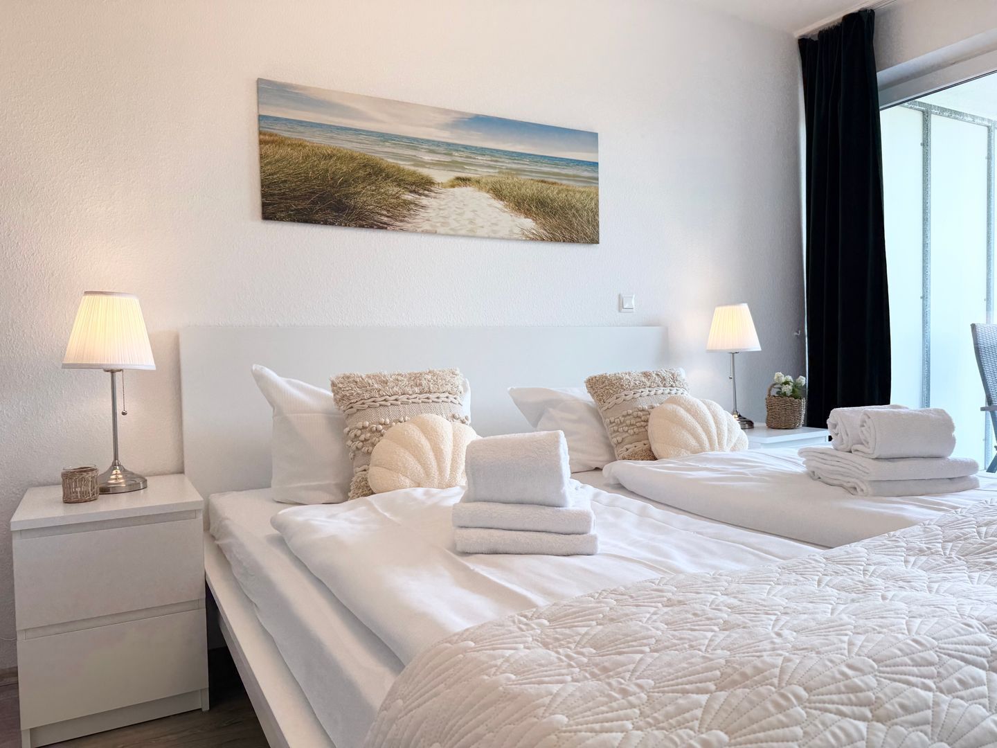 Ein Hotelzimmer mit zwei Betten, einem Strandgemälde und einem mit Vorhängen versehenen Fenster.