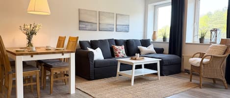 A modern living room with a sofa, coffee table, dining table, and chairs. There are three paintings on the wall, a window with curtains, and a wicker chair.