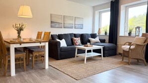Een moderne woonkamer met een bank, salontafel, eettafel en stoelen. Er hangen drie schilderijen aan de muur, een raam met gordijnen en een rieten fauteuil.