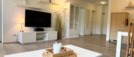 Una sala de estar moderna con televisor de pantalla plana, muebles blancos, una mesa de madera con objetos decorativos y una zona de cocina al fondo.